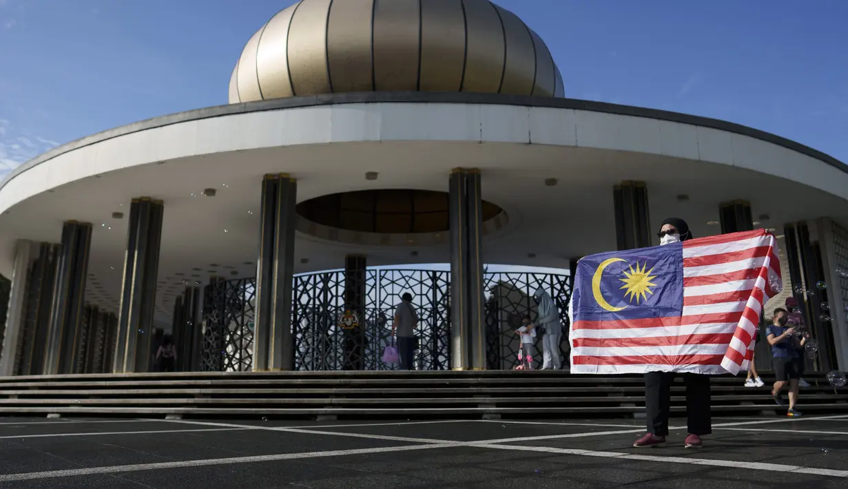 Seorang pengunjung berpose dengan bendera nasional Malaysia di depan Monumen Nasional saat perayaan Hari Nasional ke-64 untuk memperingati kemerdekaan Malaysia dari penjajahan Inggris di Kuala Lumpur, Malaysia, Selasa (31/8/2021). (AP Photo/Vincent Thian)
