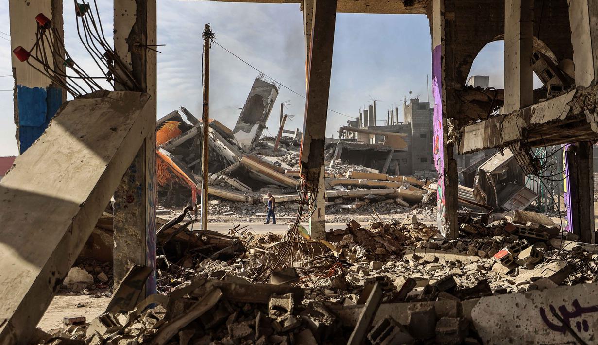 Seorang pria berjalan melewati bangunan-bangunan yang hancur yang sebagian tertutup mural di kamp pengungsi Maghazi di luar Deir al-Balah pada Senin 8 Desember 2025. Mesir meminta penempatan pasukan stabilisasi internasional secepatnya di Jalur Gaza untuk memverifikasi kesepakatan gencatan senjata antara Israel dan Hamas. (EYAD BABA/AFP)