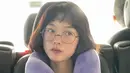Penampilannya semakin cute dengan penggunaan kacamata frame bulat. Makeup natural yang digunakan juga memperlihatkan wajahnya yang awet muda bak masih ABG. Bergaya dengan pose berpikir dan mengenakan bantal leher ungu membuatnya jadi terlihat begitu manis. (Liputan6.com/IG/@leeyoum262)