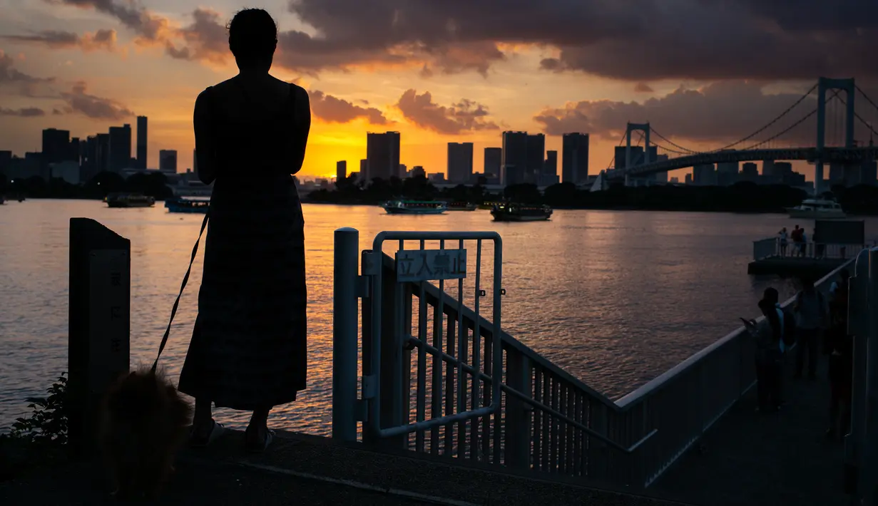 Seorang wanita bersama anjingnya menikmati matahari terbenam di Odaiba, Tokyo, (29/7). Odaiba adalah sebuah pulau buatan di Teluk Tokyo, Jepang yang dihubungkan dengan pusat kota Tokyo dengan Jembatan Rainbow. (AFP Photo/Martin Bureau)