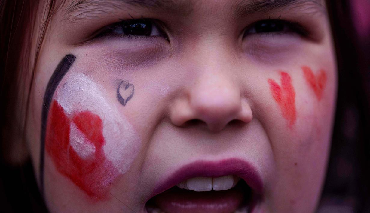 Bendera merah-putih Greenland berkibar di antara sejumlah poster penolakan. Tampak dalam foto, seorang gadis berteriak saat protes menentang kebijakan Presiden Amerika Serikat (AS), Donald Trump, terhadap Greenland di depan konsulat AS di Nuuk, Sabtu 17 Januari 2026. (AP Photo/Evgeniy Maloletka)