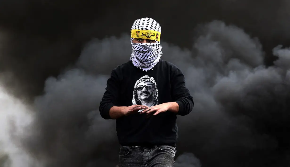 Seorang pemuda Palestina mengenakan kemeja bergambar mendiang pemimpin Yasser Arafat berjalan melewati ban yang terbakar saat konfrontasi dengan pasukan keamanan Israel menyusul demonstrasi menentang perampasan tanah oleh Israel di Desa Kfar Qaddum, Tepi Barat, 4 Maret 2022. (JAAFAR ASHTIYEH/AFP)
