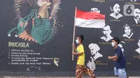 Anak-anak melintas membawa bendera merah putih berlatar belakang mural Pancasila di Kampung Pancasila, Karang Tengah, Kota Tangerang, Selasa (1/6/2021). Sejumlah kegiatan diadakan warga, Komunitas Taman Potret dan TNI ini untuk memperingati Hari kelahiran Pancasila. (Liputan6.com/Angga Yuniar)