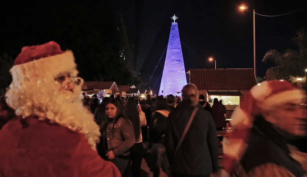 Pria berkostum Sinterklas terlihat sekitar pohon natal dari botol plastik di kota Chekka, Lebanon, 15 Desember 2019. Caroline Chaptini, yang mengorganisasi proyek ini menyebut, kreasi ini terjadi berkat kerjasama dengan pemerintah kota setempat. (Ibrahim CHALHOUB/AFP)