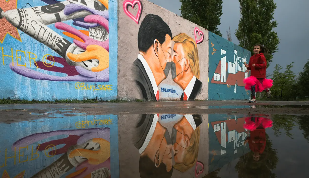 Seorang gadis berlari di depan Graffiti yang menggambarkan Presiden AS Trump (kanan) dan Presiden Tiongkok Xi Jinping saling berciuman dengan mengenakan masker di tembok taman umum Mauerpark di Berlin, Jerman, Rabu, (29/4/2020). (AP/Markus Schreiber)
