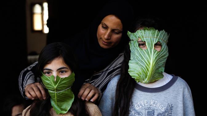 Seorang ibu memakaikan anak-anaknya masker dari kubis saat sedang memasak di rumahnya di tengah wabah pandemi corona (Covid-19) di Beit Lahia di Jalur Gaza utara (16/4/2020). Hal itu dilakukan agar anak-anaknya tak cemas di tengah wabah pandemi. (AFP/Mohamed Abed)