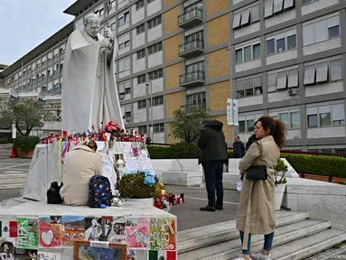 Orang-orang berdiri di depan patung Yohanes Paulus II di luar Rumah Sakit Universitas Gemelli tempat Paus Fransiskus dirawat karena pneumonia, di Roma pada 8 Maret 2025. (ANDREAS SOLARO/AFP atau pemberi lisensi/AFP)