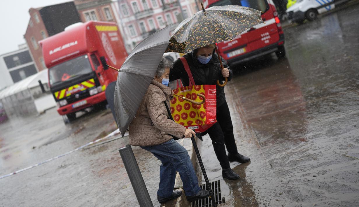 Seorang perempuan membantu seorang lainnya menyeberang jalan di tengah hujan, di samping kendaraan perlindungan sipil yang beberapa jalan terendam banjir, di Alges, tepat di luar Lisbon, Selasa (13/12/2022). Badai Atlantik menghantam semenanjung Iberia, meninggalkan jejak kehancuran pada Selasa, terutama di Ibu kota Portugal,  Lisbon, sebelum pindah ke timur ke Spanyol.  (AP Photo/Armando Franca)