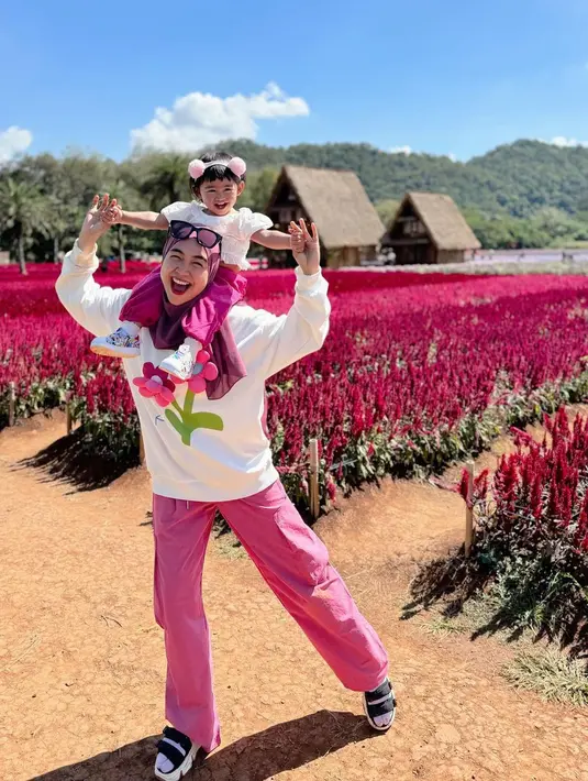 Gaya ceria berlatar kebun bunga, Ria Ricis berpose gemas dengan Moana. Ricis mengenakan sweater putih bercorak bunga berwarna ungu yang serasi dengan cargo pants pinknya, menggendong Moana di atas pundaknya, yang mengenakan atasan lengan pendek berwarna putih dan celana pink yang serasi. [Foto: Instagram/riaricis1795]