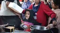 Kantor media pemerintah Gaza dalam pernyataannya pada Minggu 12 April 2026, menyebut kondisi saat ini sebagai dampak pembatasan pasokan yang terus berlangsung oleh Israel. Tampak dalam foto, anak-anak pengungsi Palestina menerima makanan dari dapur amal di kamp pengungsi Nuseirat, Jalur Gaza Tengah pada 13 April 2026. (Eyad Baba/AFP)
