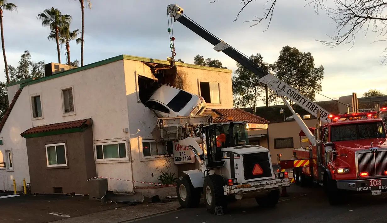 Petugas mengevakuasi mobil sedan putih yang tertancap di bangunan lantai dua klinik dokter gigi di Santa Ana, California, Minggu (14/1). Separuh badan mobil terjepit di gedung tersebut setelah melewati pembatas jalan. (Orange County Fire Authority via AP)