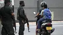 Para komuter mengendarai skuter melewati tentara yang berjaga di sepanjang jalan di Kolombo pada Sabtu (2/4/2022). Pasukan yang dipersenjatai dengan senapan serbu otomatis dikerahkan di Sri Lanka untuk mengendalikan massa, beberapa jam setelah presiden menyatakan keadaan darurat. (KODIKARA / AFP)