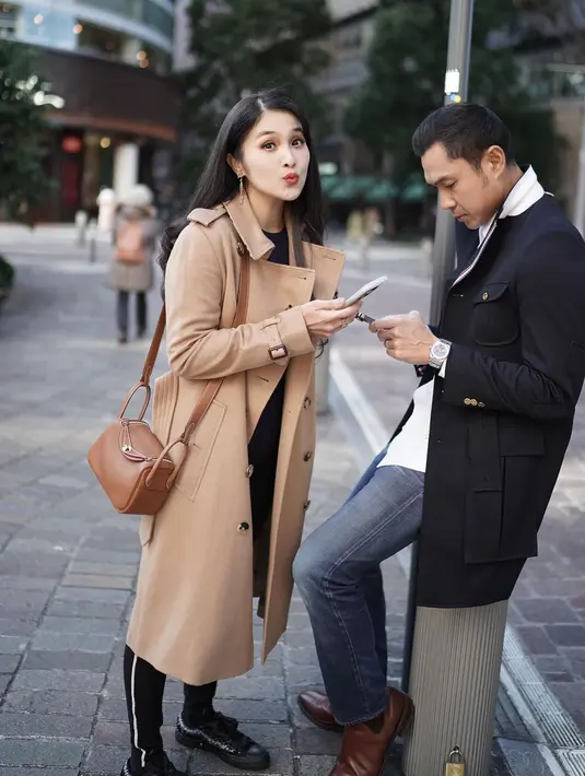 Sandra Dewi tampil dengan long coat cokelat dan celana panjang hitam yang senada dengan sepatunya. Sedangkan Harvey yang tampak sibuk dengan ponselnya tampak mengenakan kemeja putih, double-breasted long coat hitam, celana jeans, dan sepatu cokelat. Foto: Instagram.