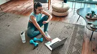 Seorang wanita sporty dalam pakaian olahraga sedang duduk di lantai dengan dumbel dan protein shake atau sebotol air dan menggunakan laptop di rumah di ruang tamu. Konsep olahraga dan rekreasi/Shutterstock-ORION PRODUCTION.
